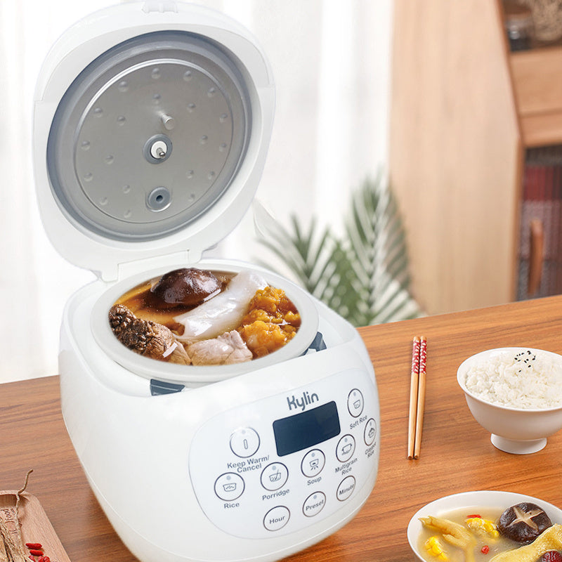 White rice cooker with food on a wooden table, surrounded by bowls and chopsticks.